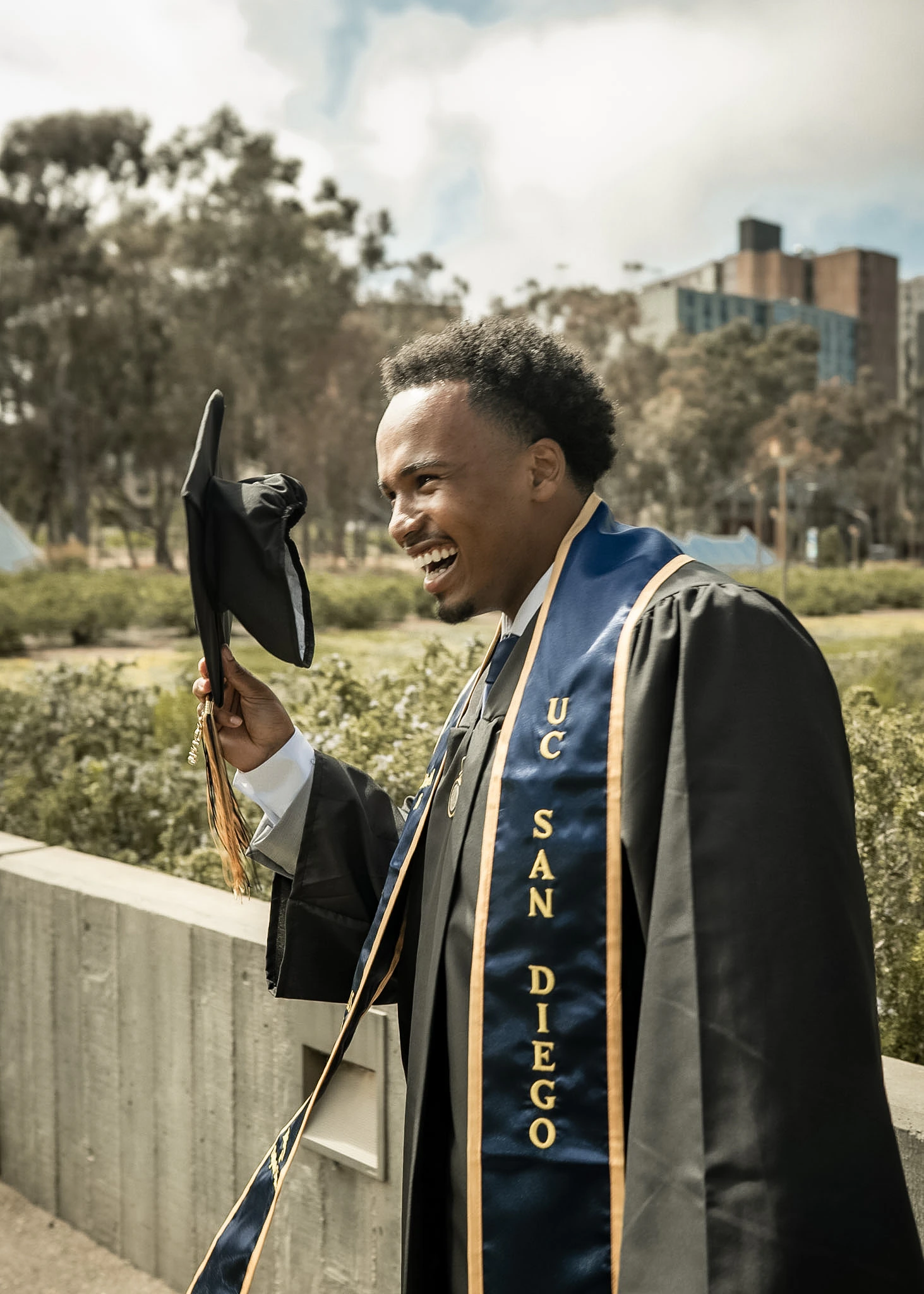 Graduate laughing outdoors while holding a commencement cap.